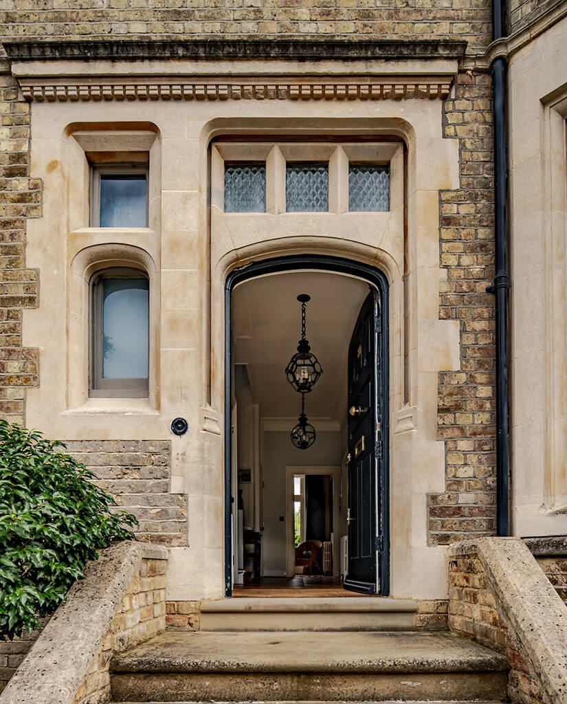 A stone arch doorway with an open dark door leads into a hallway with pendant lights in a brick building. Steps and a bush are in the foreground.
