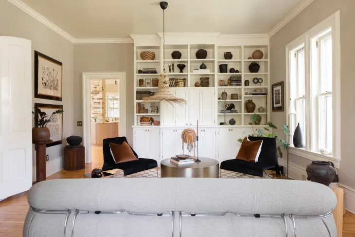 Contemporary living room with a white built-in bookshelf, two black chairs, a round table, and a pendant light. Large windows let in natural light, and the decor is neutral and modern.