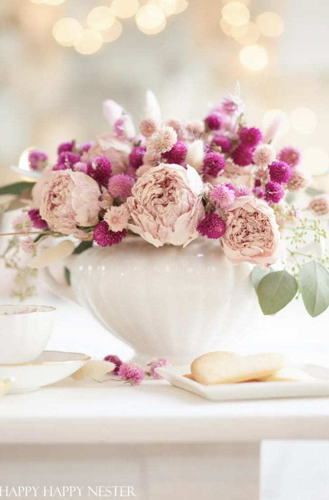 A white ceramic vase holds a bouquet of pale pink roses and small purple flowers, placed on a white table next to a plate with cookies and a teacup.