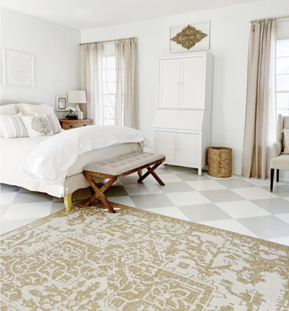 Bedroom with white walls and furniture, beige curtains, patterned rug, wooden bench at the bed’s foot, and natural light from two windows.