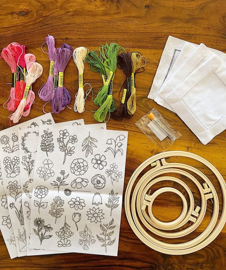 Embroidery kit with colored threads, four fabric squares, three embroidery hoops, two needles in a case, and floral pattern sheets on a wooden surface.