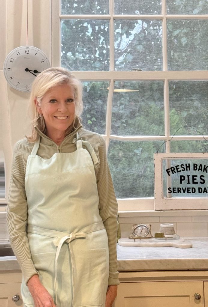 A woman wearing an apron stands in a kitchen in front of a window, with a clock and a sign reading "Fresh Baked Pies Served Daily" visible in the background.