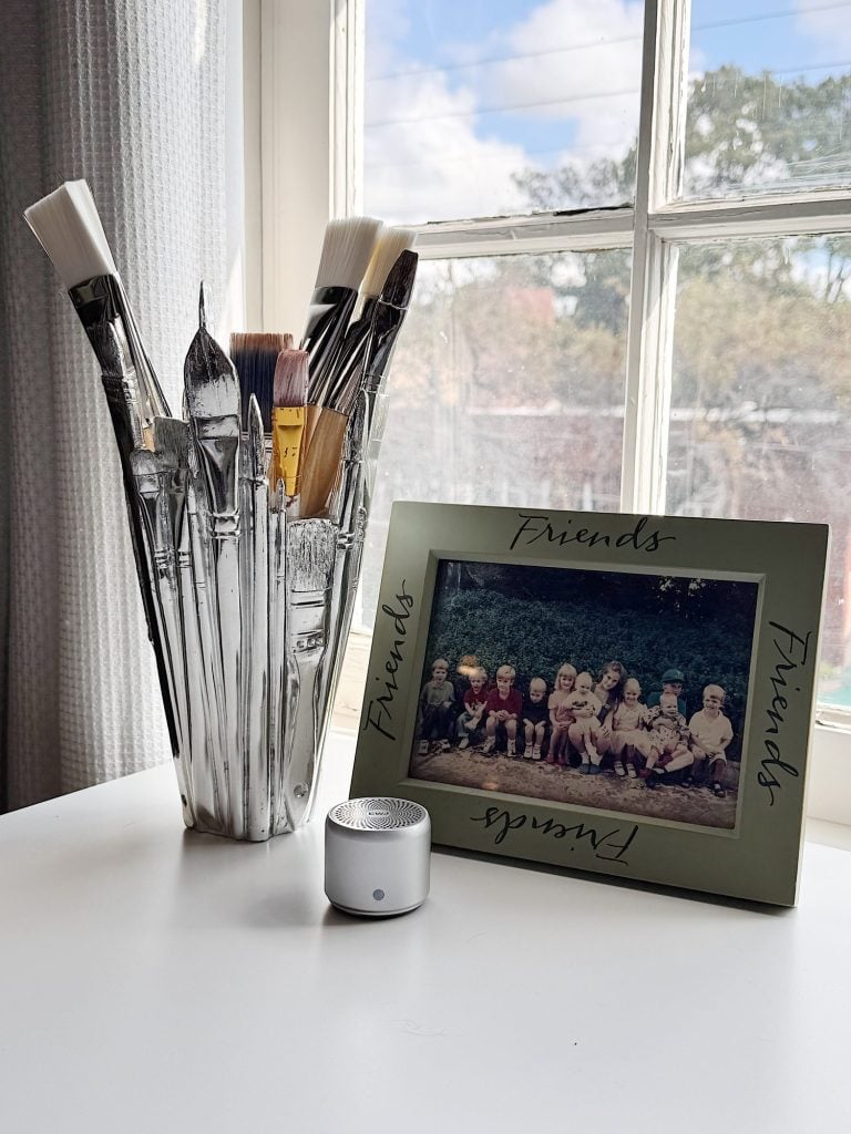 A glass holds paintbrushes next to a framed group photo labeled "Friends" and a small speaker, all on a white table by a window.