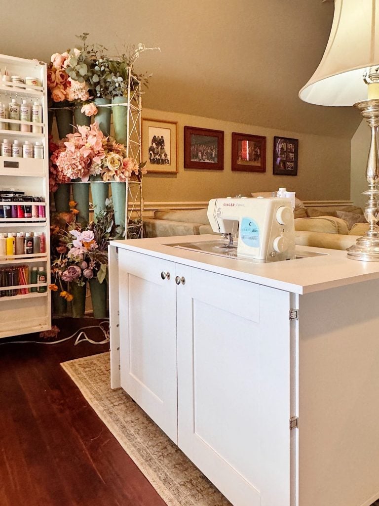 A sewing machine sits on a white cabinet in a craft room, with shelves of paint, artificial flowers, and framed pictures on the wall.