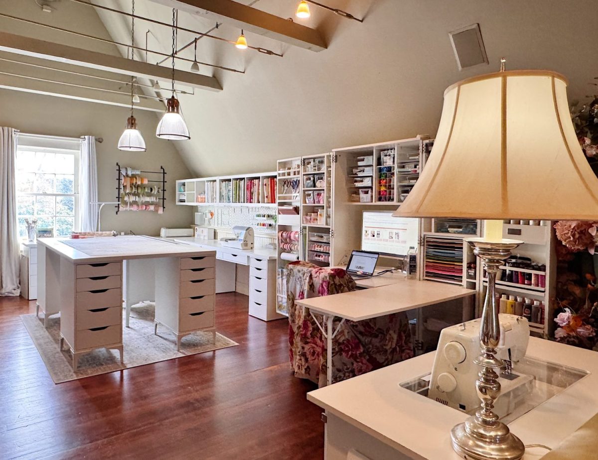 A spacious, well-lit craft room with organized shelves of supplies, two large work tables, a desk with a laptop, and a sewing machine next to a lamp.