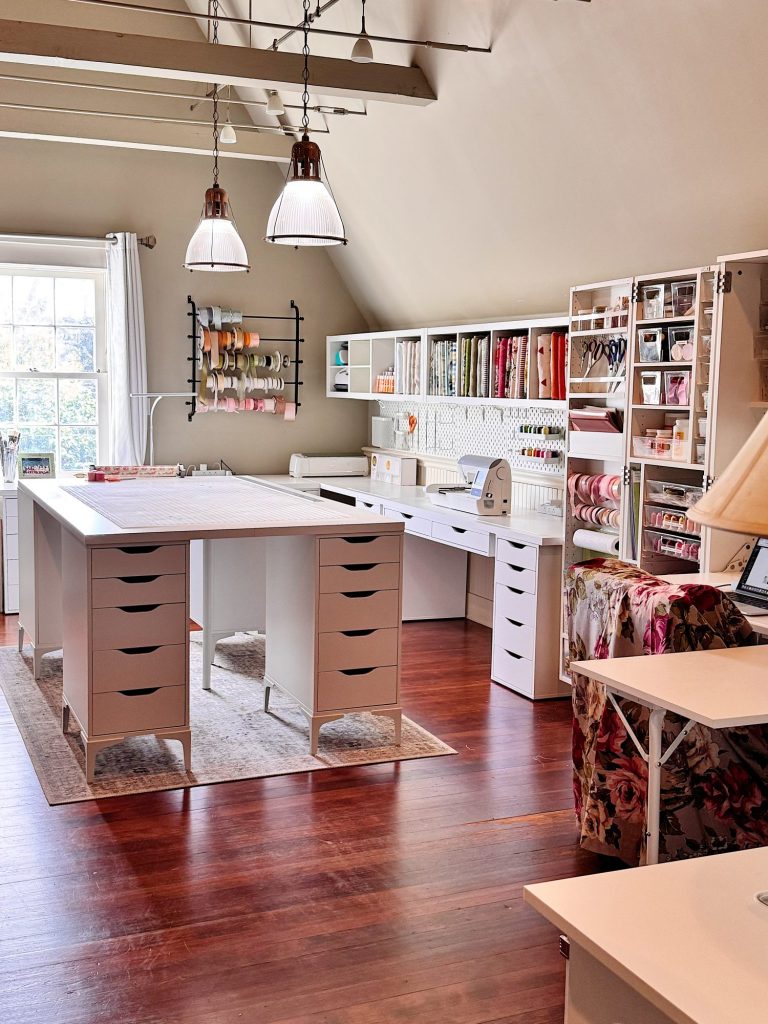 A spacious, organized craft room with large worktables, storage drawers, fabric rolls, and supplies neatly arranged on shelves and wall racks. Natural light comes in through a window.