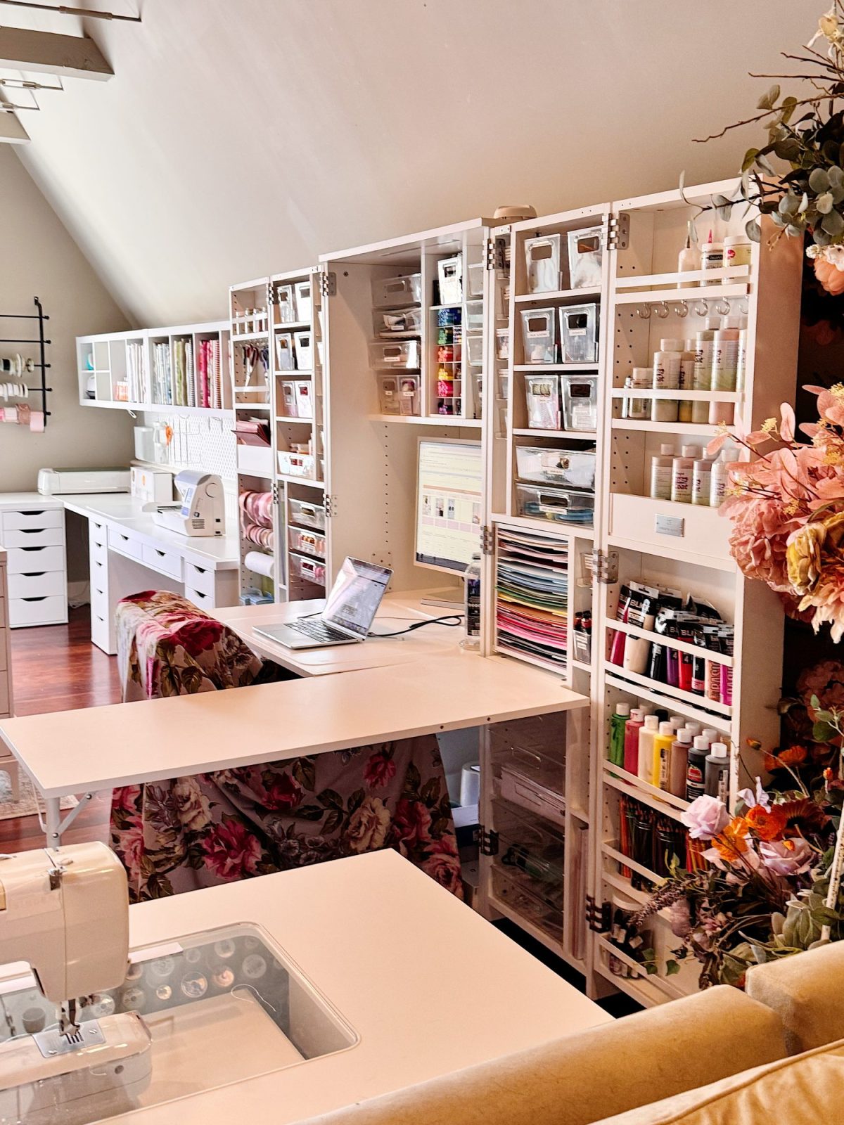 Well-organized craft room with white shelving units, various art supplies, a laptop, and a sewing machine, featuring ample storage and floral decorations.