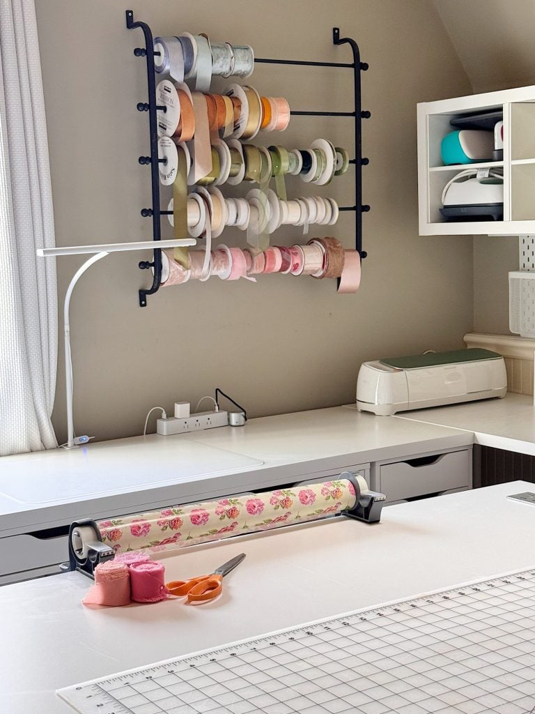 A craft room workspace features a wall-mounted ribbon rack, a cutting mat, scissors, tape, floral wrapping paper, and a cutting machine on a white countertop.