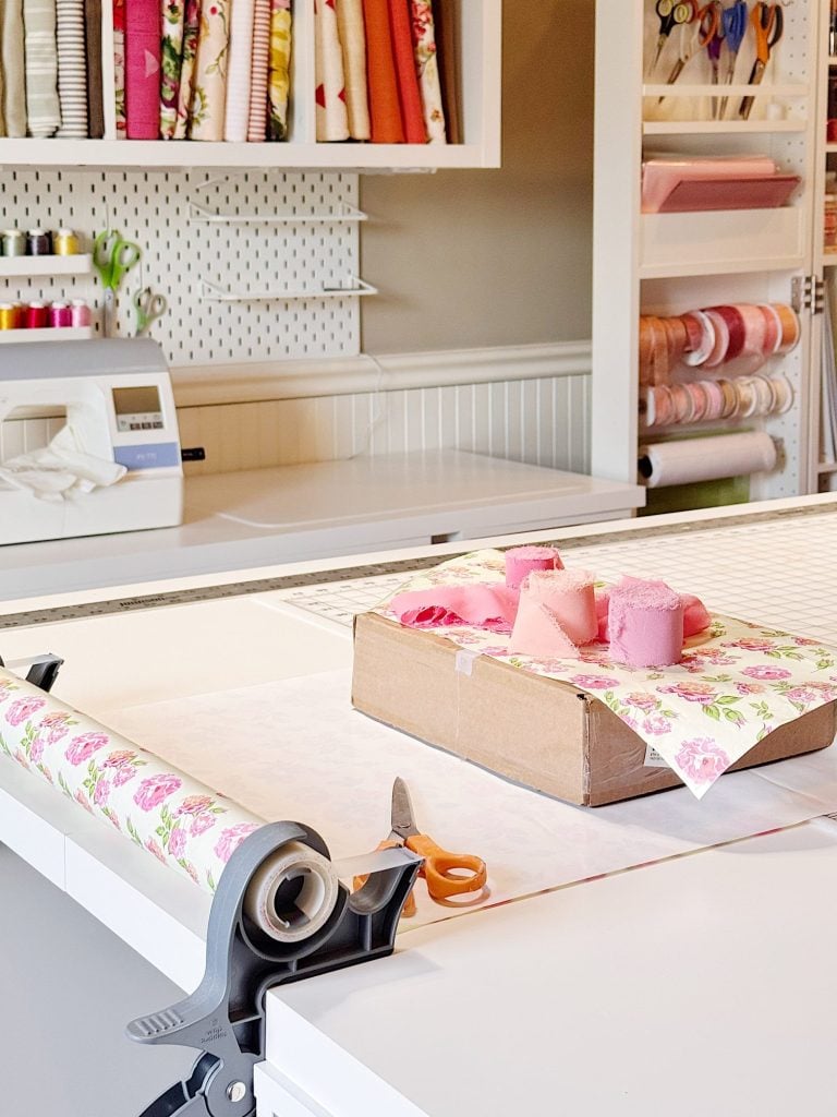 A fabric cutting station with a box, pink and floral fabrics, scissors, a rotary cutter, and organized sewing supplies in the background.