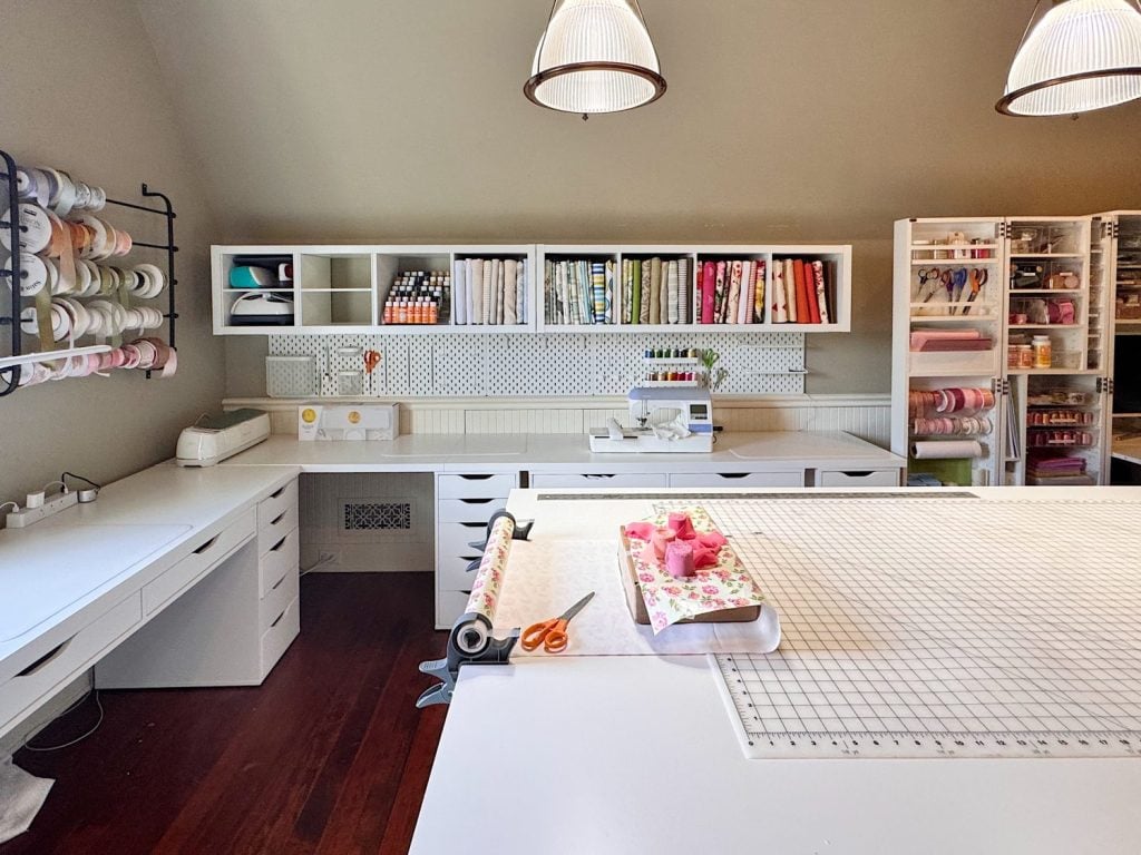 A well-organized craft room with white desks, shelving holding fabric, ribbons, and supplies, sewing machines, and a large cutting mat on the central table.