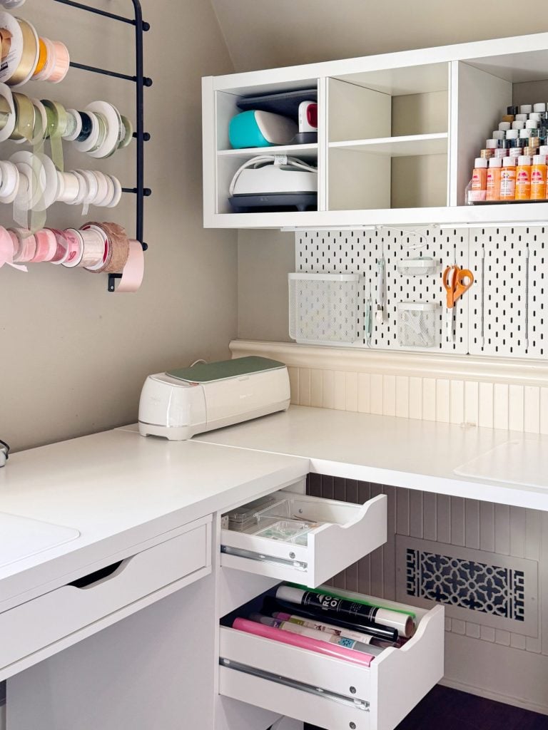 A neatly organized craft room with white shelves, a cutting machine, rolls of ribbon, bottles of paint, scissors, and open drawers containing crafting supplies.