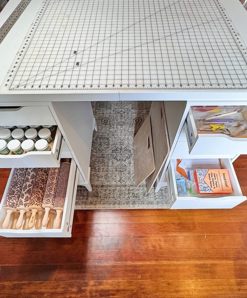 Overhead view of a craft table with a cutting mat on top, open drawers holding paint jars, patterned rolling pins, and craft supplies, on a rug and wood floor.