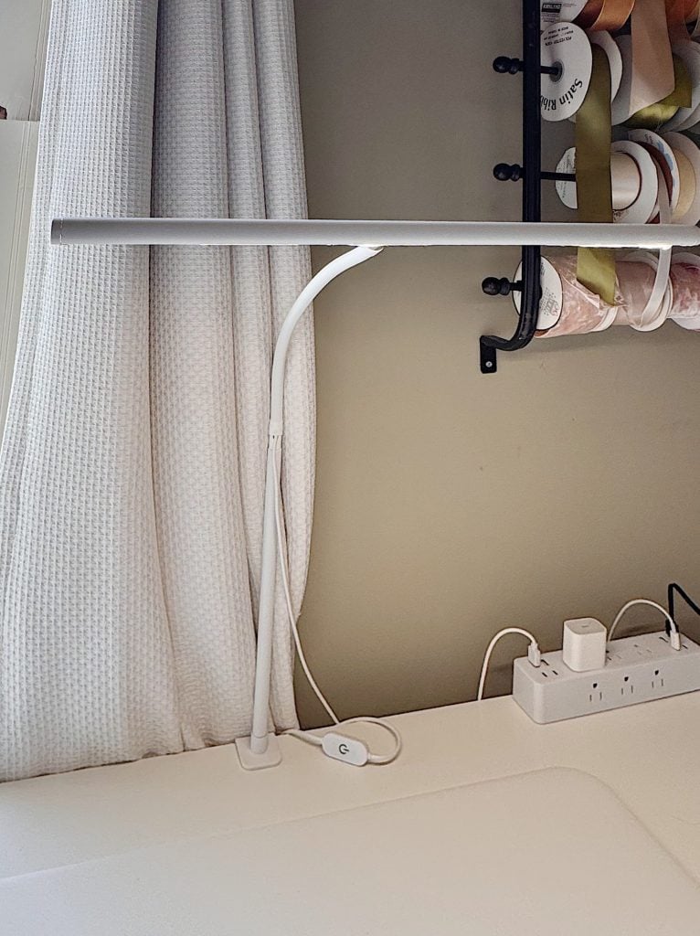 A white desk lamp with a slim horizontal light bar sits on a desk next to a power strip and a rack holding spools of ribbon.