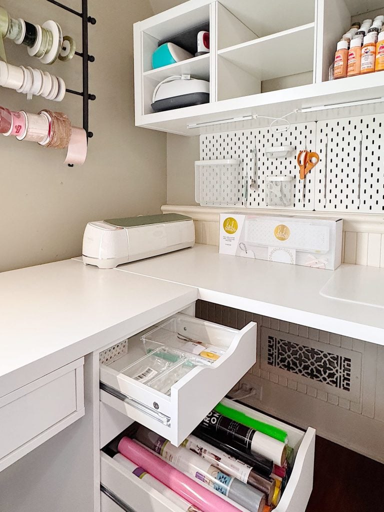 A white craft workspace with organized drawers containing vinyl rolls, a Cricut machine on the counter, and shelves holding craft supplies and ribbons.