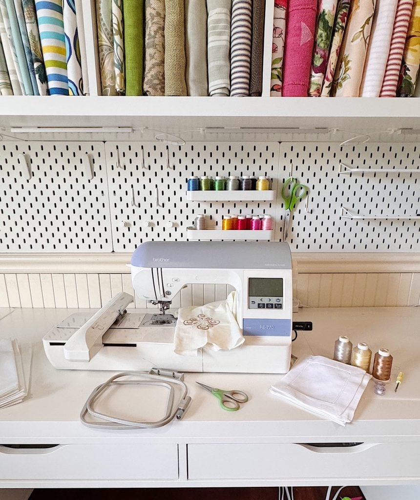 A sewing machine on a white desk with thread spools, scissors, fabric, and embroidery supplies; fabric rolls and colored threads are organized on shelves above.