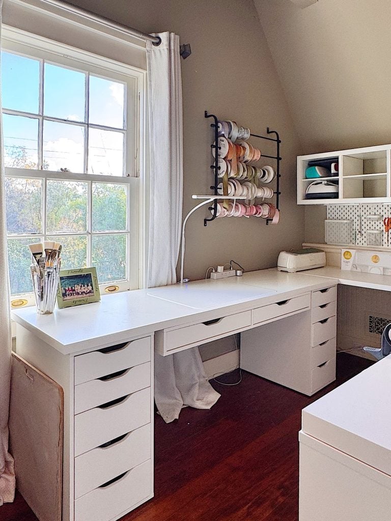 A bright, organized craft room with a white desk, drawers, storage shelves, ribbon rack, and a large window letting in natural light.