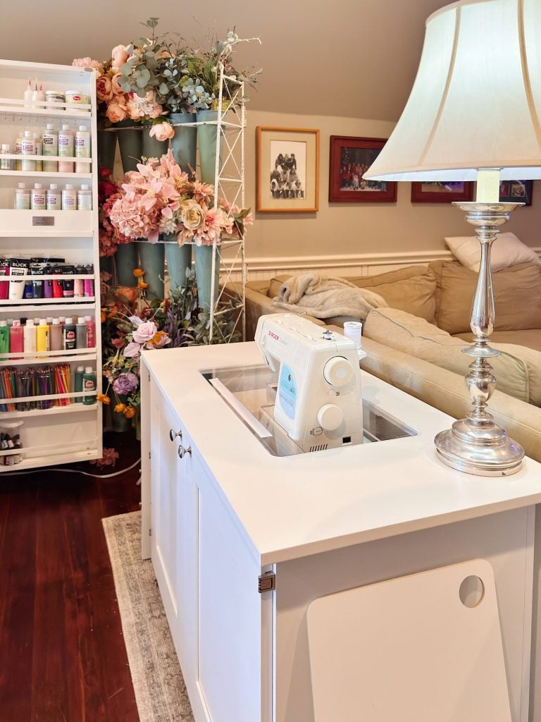 A craft room with a white sewing table, a sewing machine, a lamp, shelves of paints, and decorative flowers in vases.