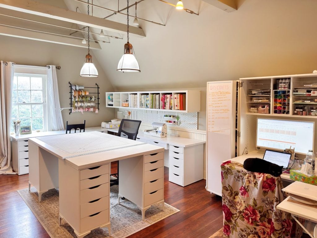 A well-organized craft room with white desks, storage drawers, shelves with supplies, a central work table, a computer, and a window letting in natural light.