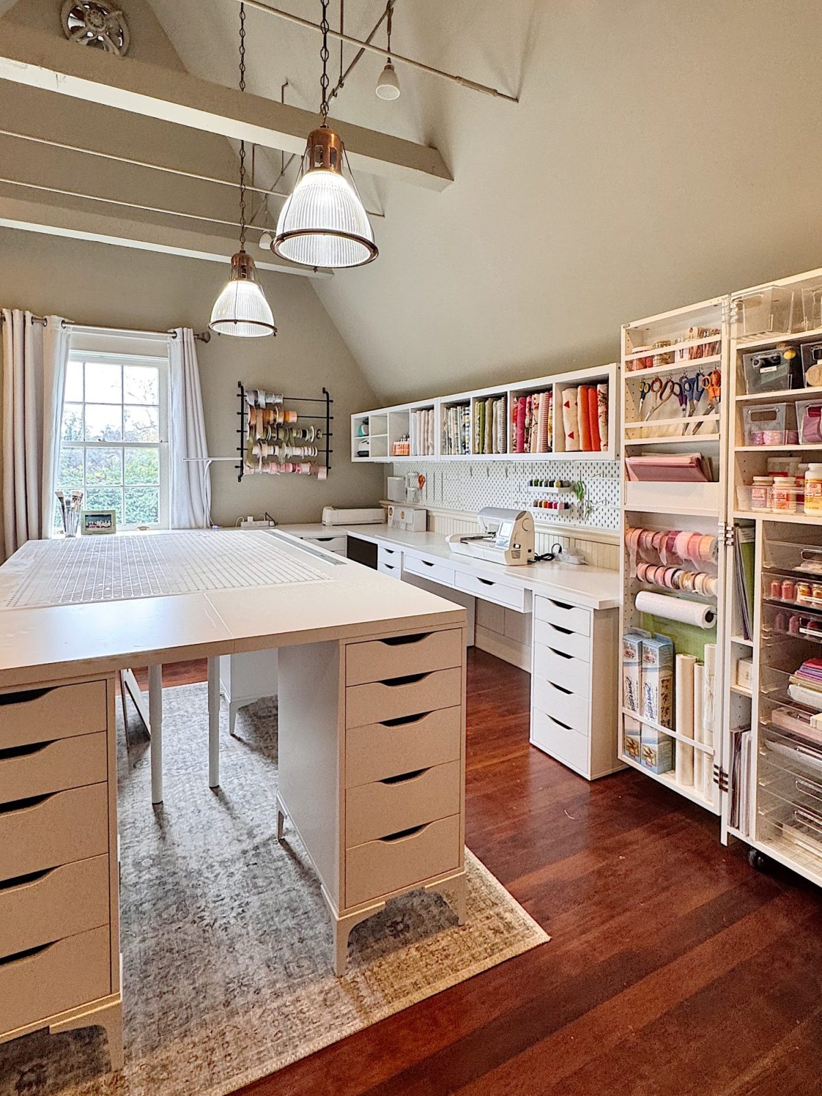 A well-organized craft room with large work tables, storage shelves filled with supplies, and natural light from a window.