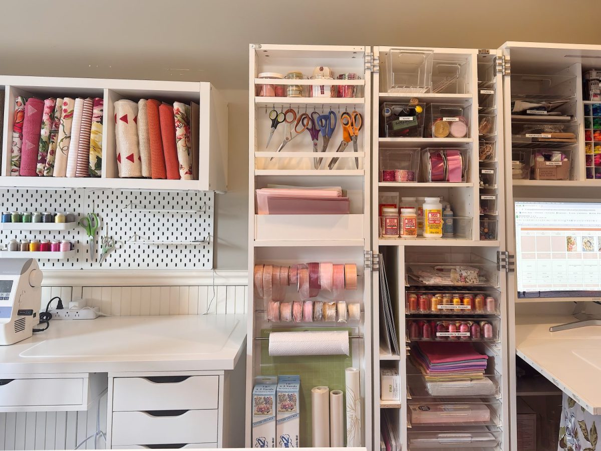 A well-organized craft room with shelves holding fabric, ribbons, scissors, craft supplies, and a computer monitor on the right side.