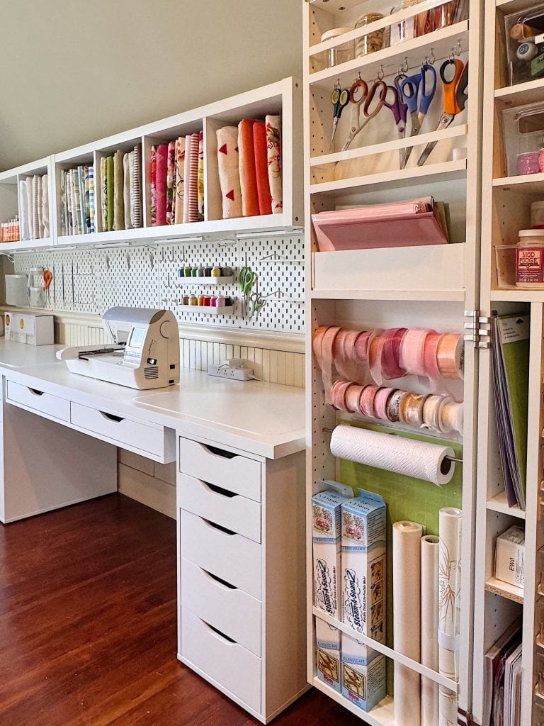 A well-organized craft room with white shelves, drawers, a desk, rolls of fabric, ribbon, scissors, and other crafting supplies neatly arranged.