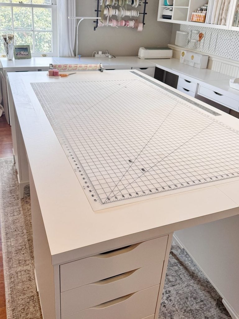 A white craft table with a large clear cutting mat sits in a well-organized workspace with drawers, shelves, and crafting supplies in the background.