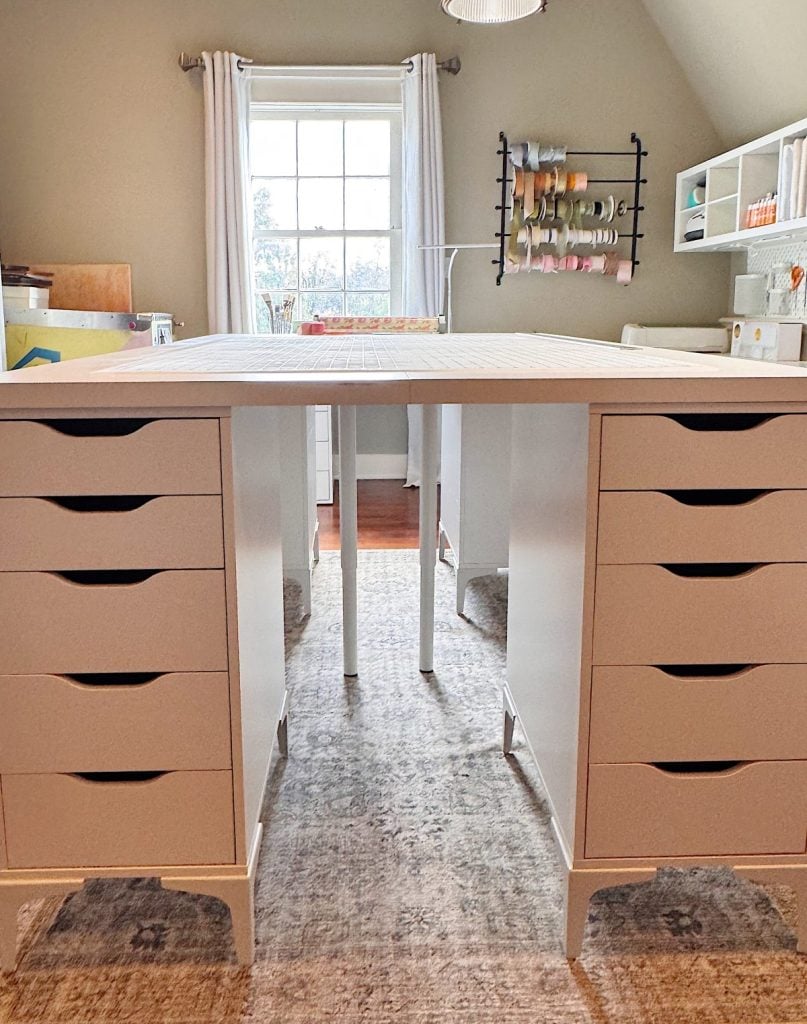 A craft room with a large work table made of two drawer units, a window with curtains, shelves, and a wall-mounted thread rack in the background.