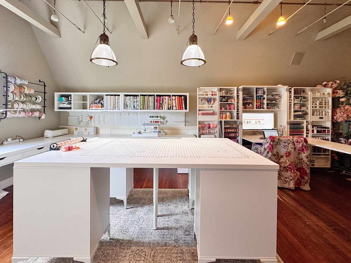 A spacious craft room with large white worktables, shelves filled with supplies, and organized storage units against the walls under pendant lights.