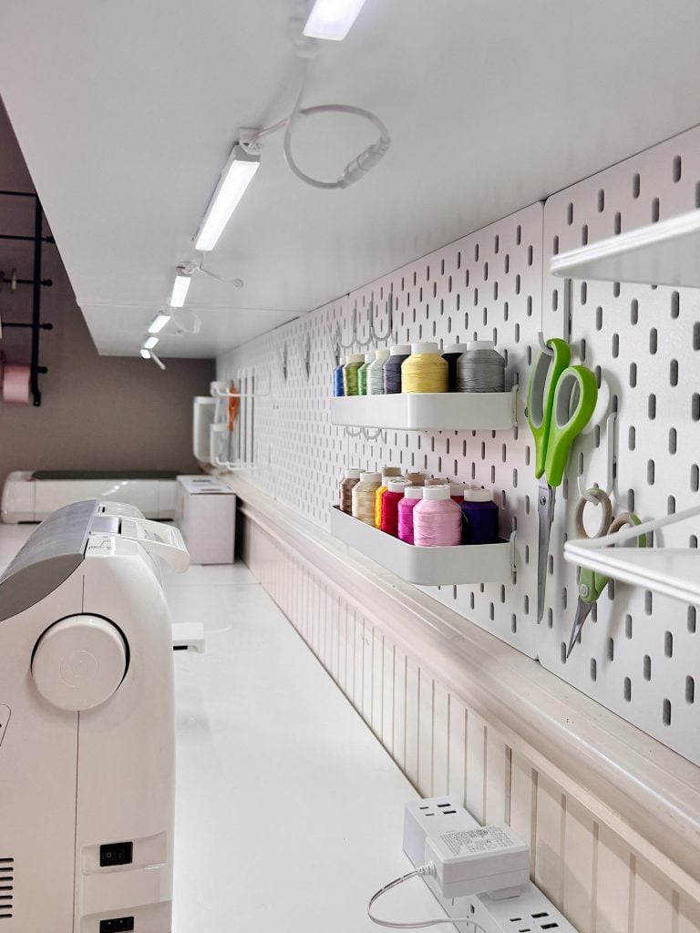 A sewing workspace with a sewing machine, thread spools, scissors, and tools organized on a white pegboard above a white countertop.