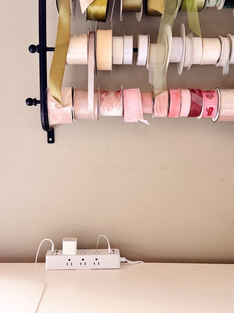 A wall-mounted rack holding various rolls of ribbon above a white desk with a power strip and charging cables against a beige wall.