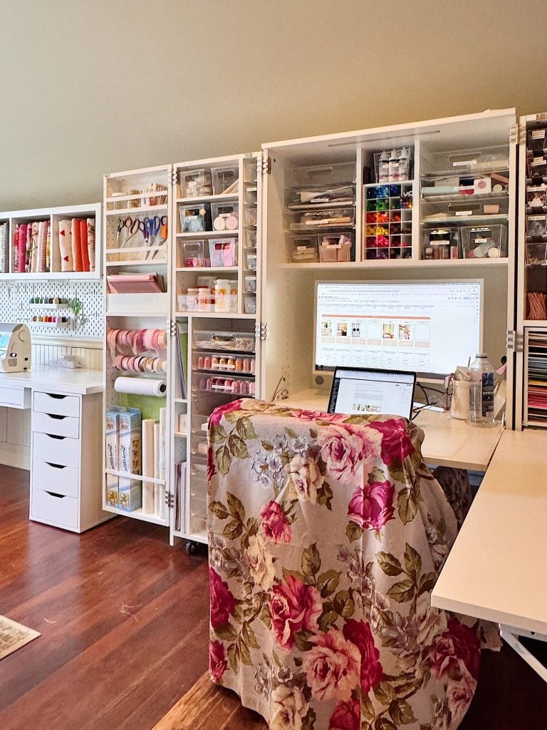 A craft room with organized shelves of supplies, a computer on a desk, and a chair covered with a floral-patterned fabric.