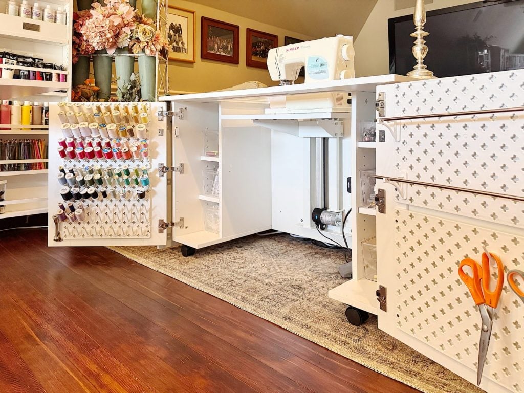 A sewing station with an open cabinet displaying thread spools, a sewing machine on a white table, and drawers with scissors on the side.