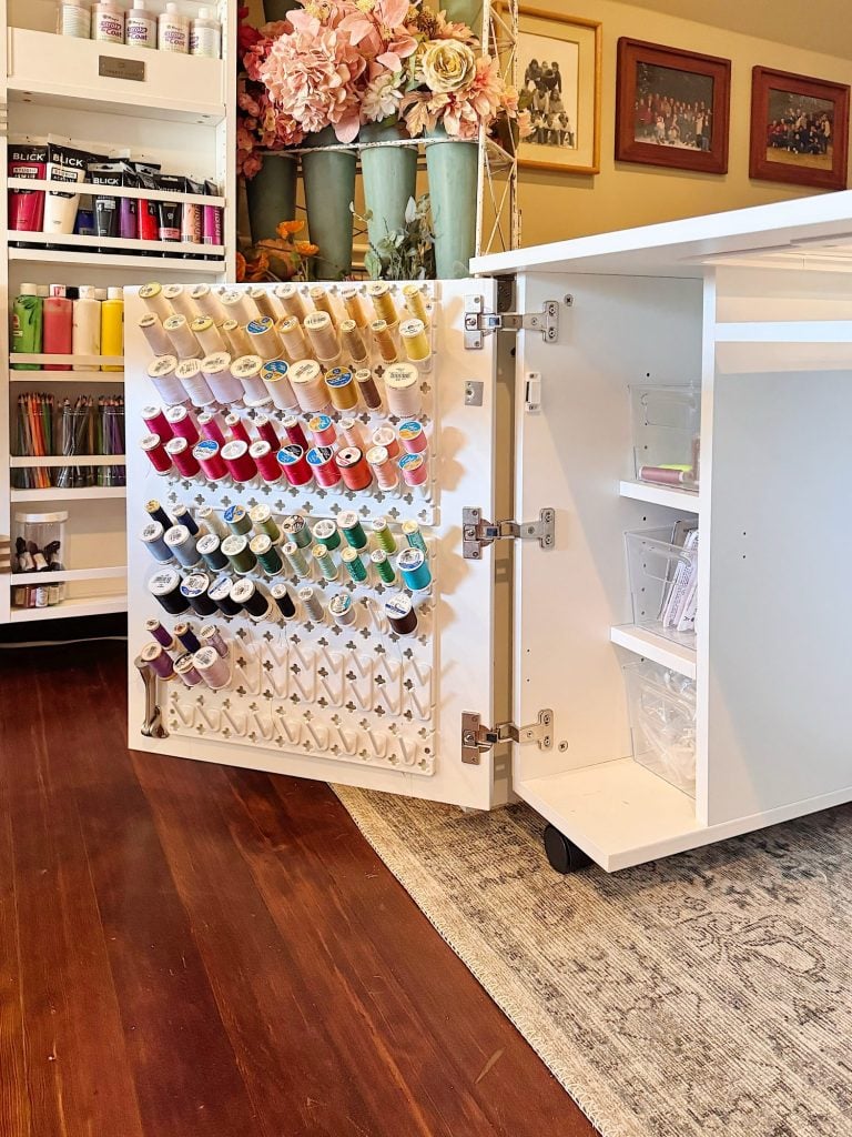 A white craft cabinet with an open door displays organized spools of thread, craft supplies, and clear storage containers. Shelves and flowers are visible in the background.