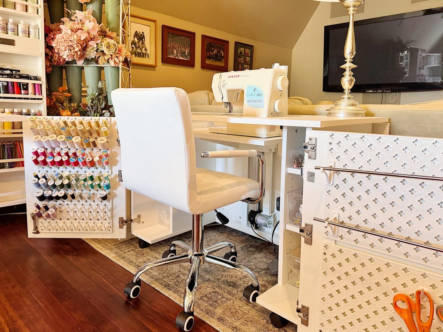 A sewing station with a white chair, sewing machine, thread rack, storage cabinets, and a lamp in a well-lit room with framed photos on the wall.