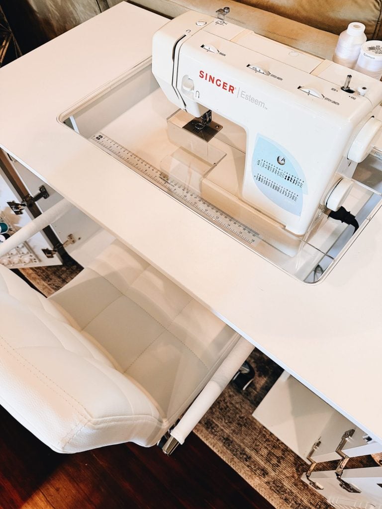 A white Singer Esteem sewing machine is set into a white sewing table with a matching white cushioned chair positioned in front of it.