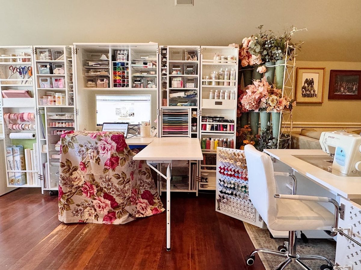 Well-organized craft room featuring shelves with supplies, a computer, sewing machines, a chair, and floral decor on wood flooring.