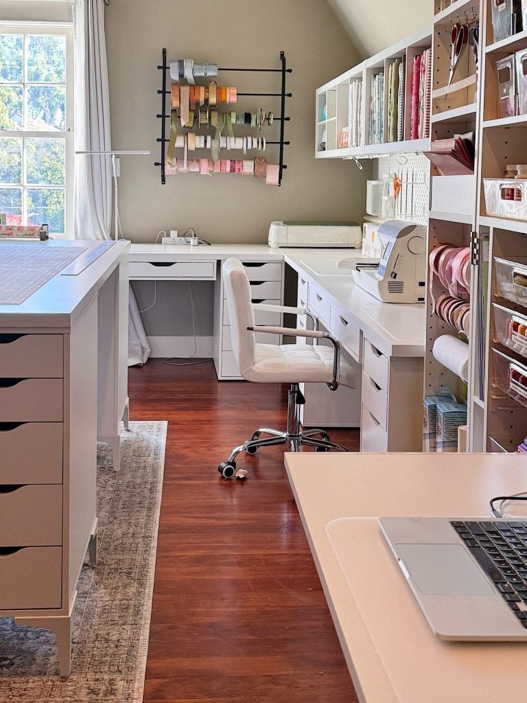A well-organized craft room with white desks, a white chair, various crafting supplies, ribbon storage on the wall, and natural light from a window.
