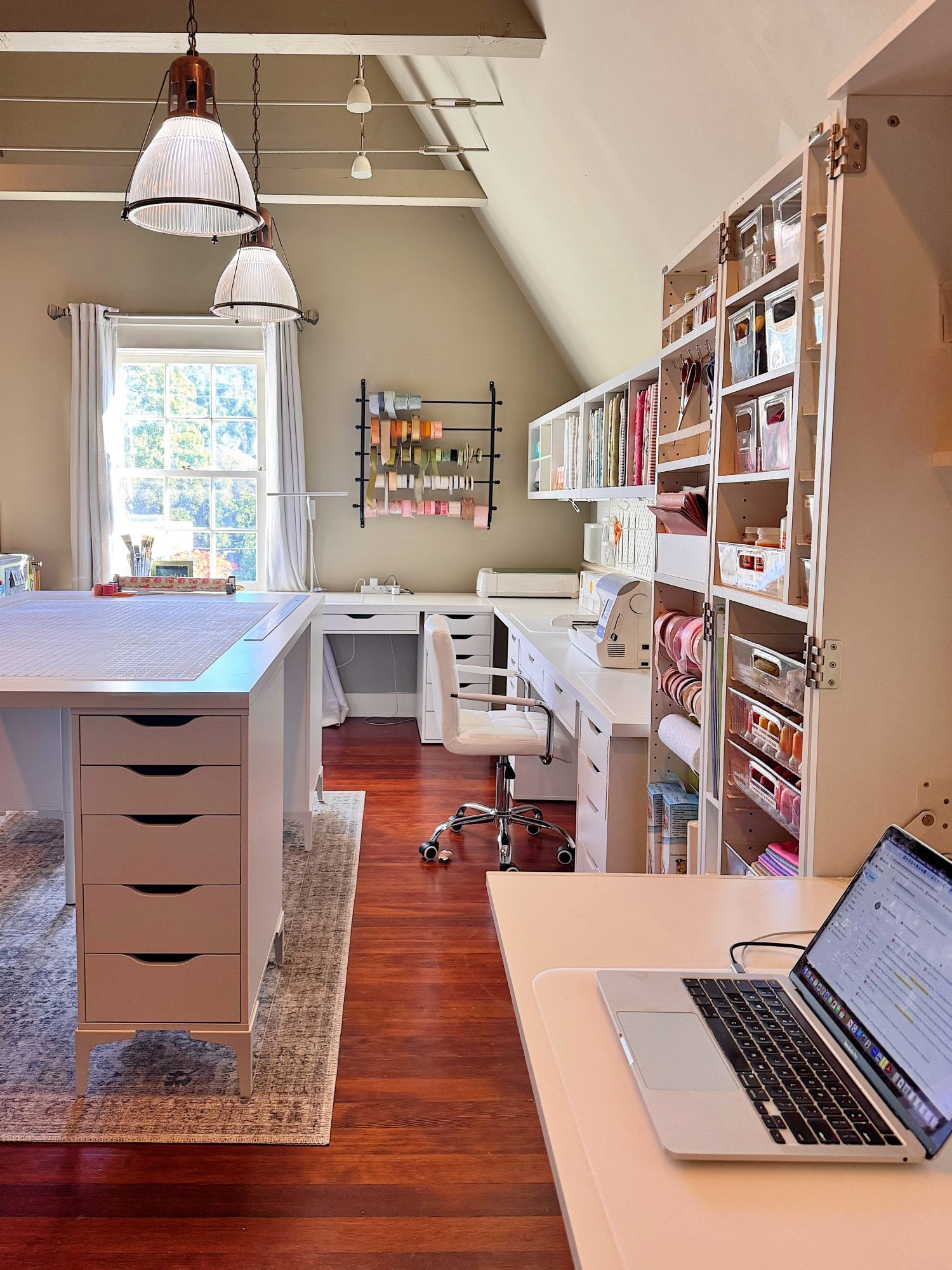 A well-organized craft room with a large worktable, shelves filled with supplies, a sewing machine, and a laptop on a desk near a window with natural light.