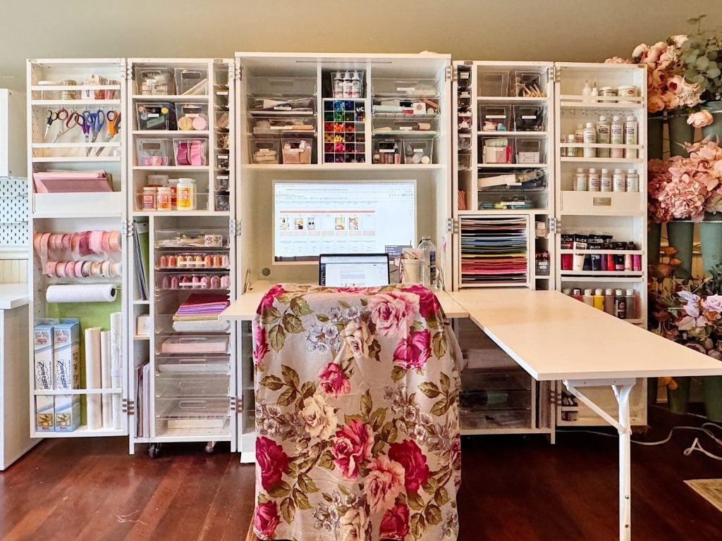Organized craft workspace with shelves holding art supplies, ribbons, and paper. A computer is on the desk, which is covered by a floral cloth. Tables are extended on both sides.