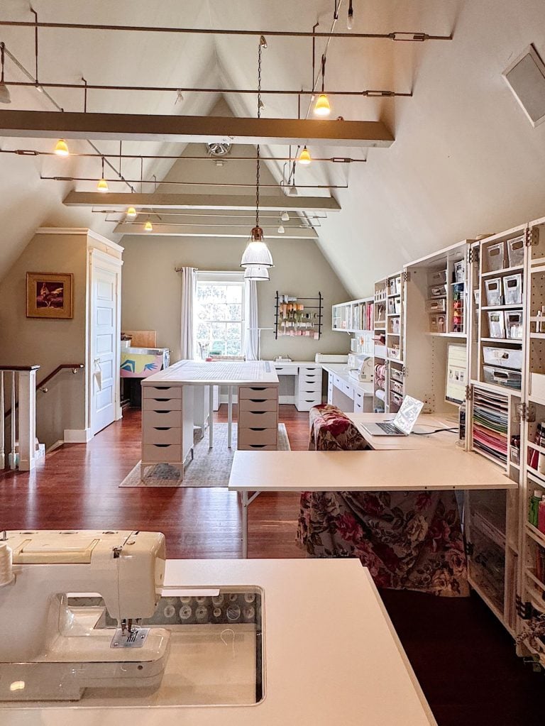 A spacious, organized craft room with large worktables, white storage drawers, wall shelves, and ample natural light from a window. A sewing machine and laptop are visible on the tables.