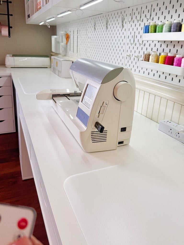 A sewing machine sits on a white countertop in a well-organized craft room with shelves holding colorful spools of thread and various supplies.