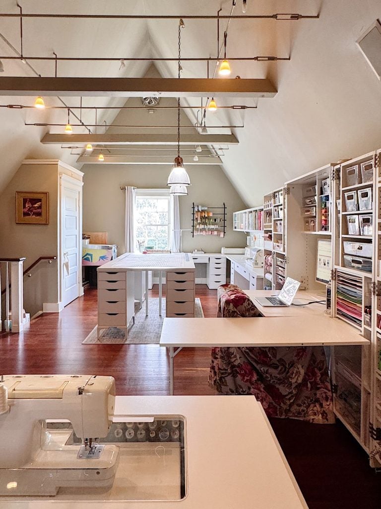 A well-organized craft room with large work tables, storage shelves, a sewing machine, a laptop, and bright natural light from a window.