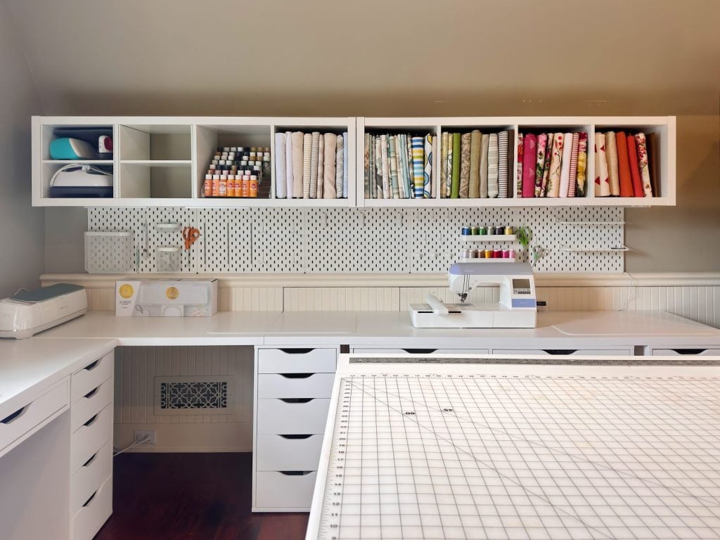 A sewing and craft room with a sewing machine, fabric rolls, organized shelves with supplies, and a large cutting mat on a white worktable.