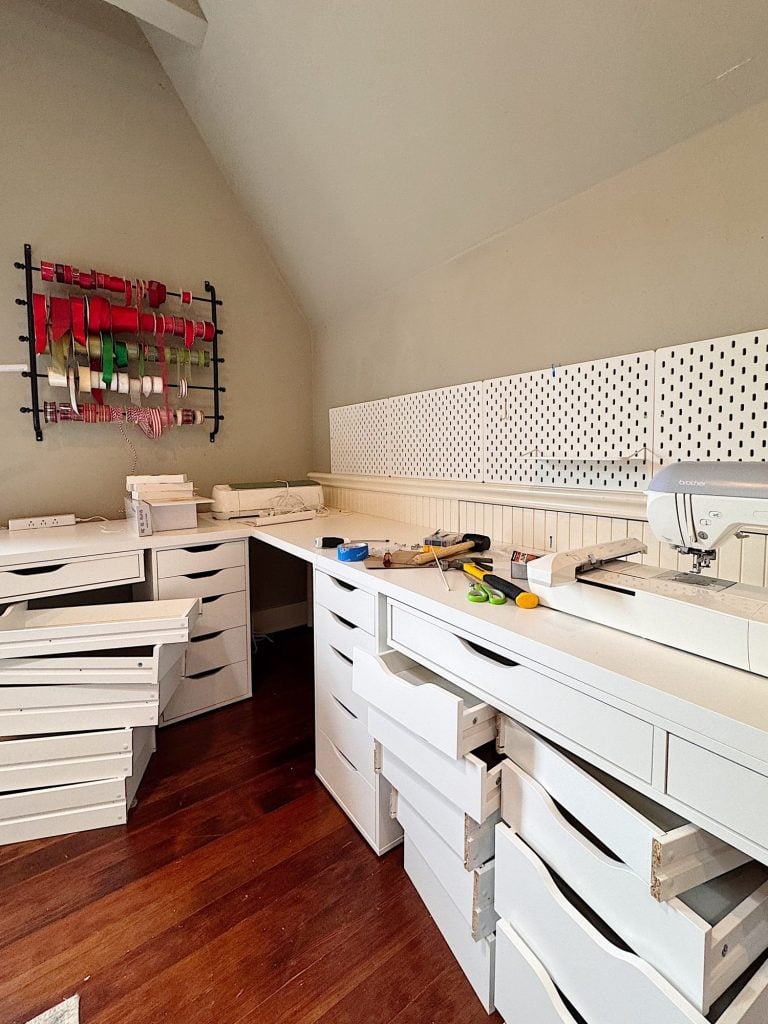 A craft room with open white drawers, tools on the countertop, a sewing machine, pegboards, and a wall rack holding rolls of ribbon.