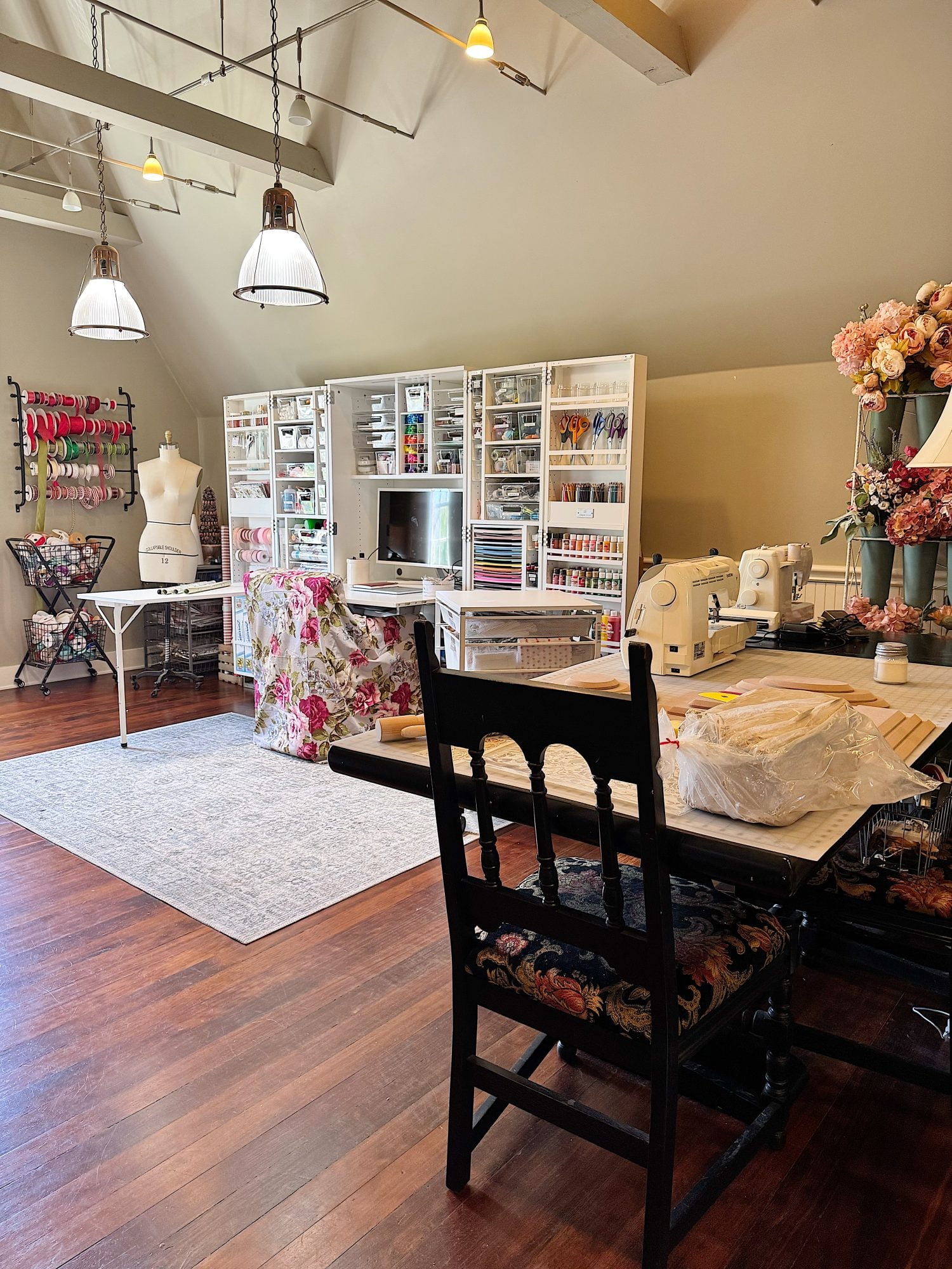 A well-organized sewing and craft room with storage cabinets, a dress form, sewing machines, work tables, and floral decor on wood flooring.