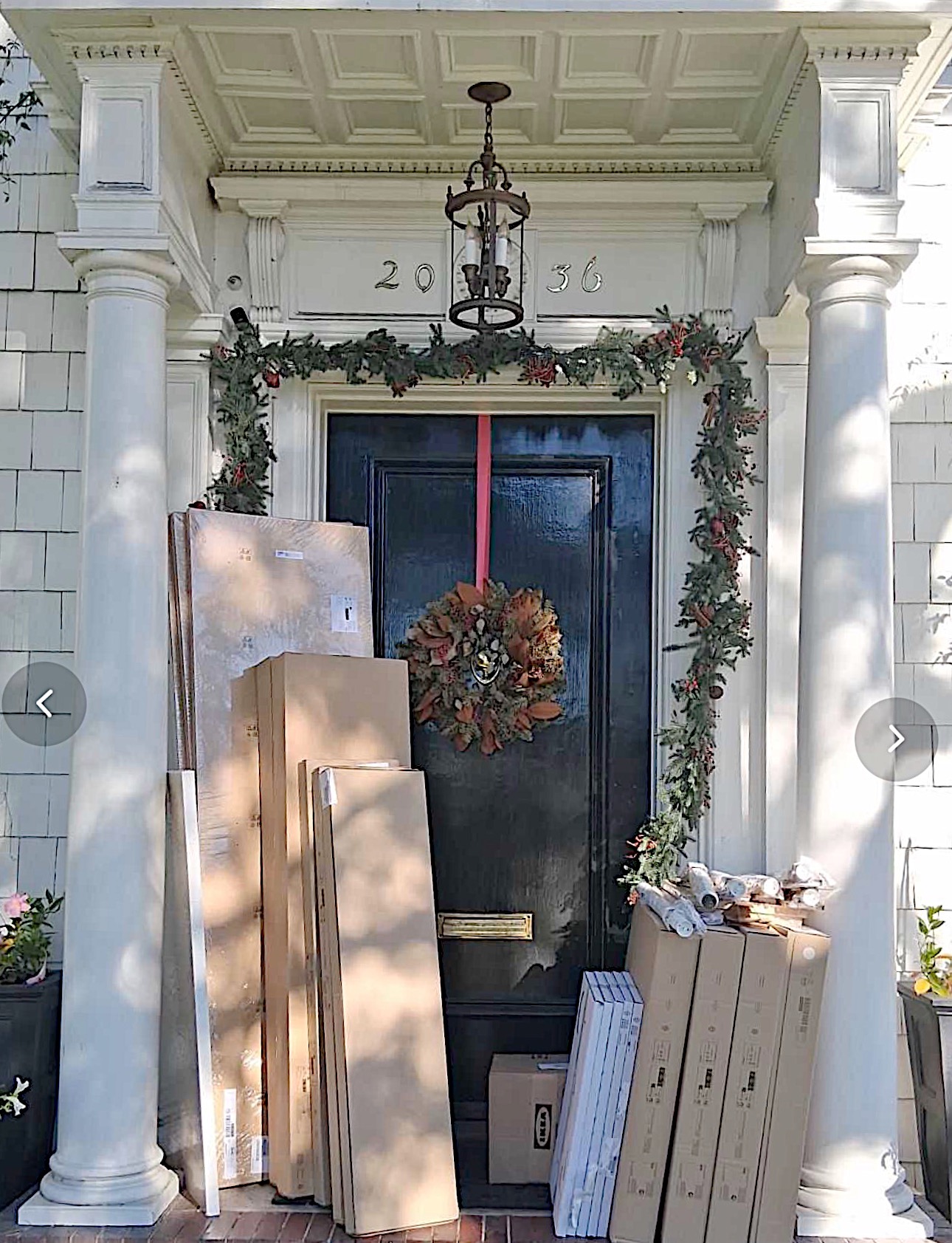 Several cardboard boxes and packages are stacked on the porch in front of a dark door decorated with a wreath and holiday garland.