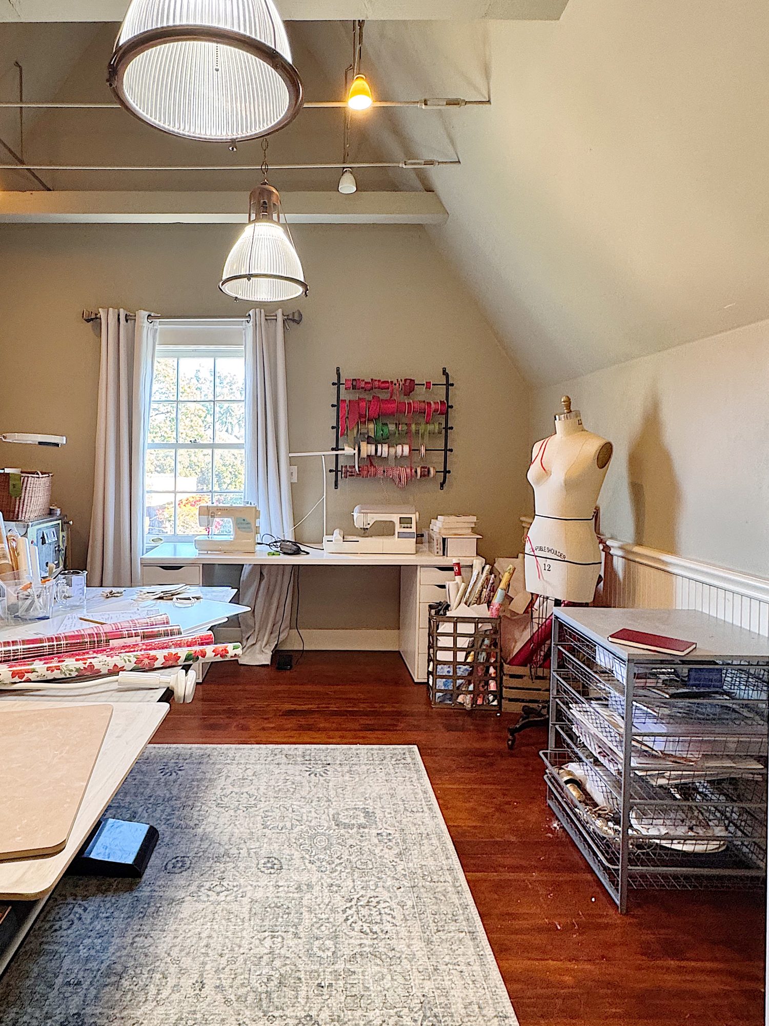 A sewing room with a dress form, sewing machine, rolls of fabric, organized shelves, and a large window letting in natural light.