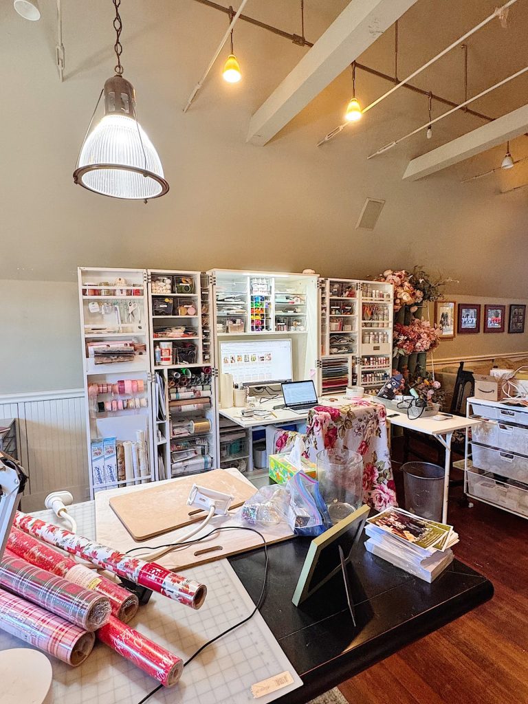 A craft room with organized shelves of supplies, a worktable with wrapping paper, tools, and a laptop, and floral decorations in the background.