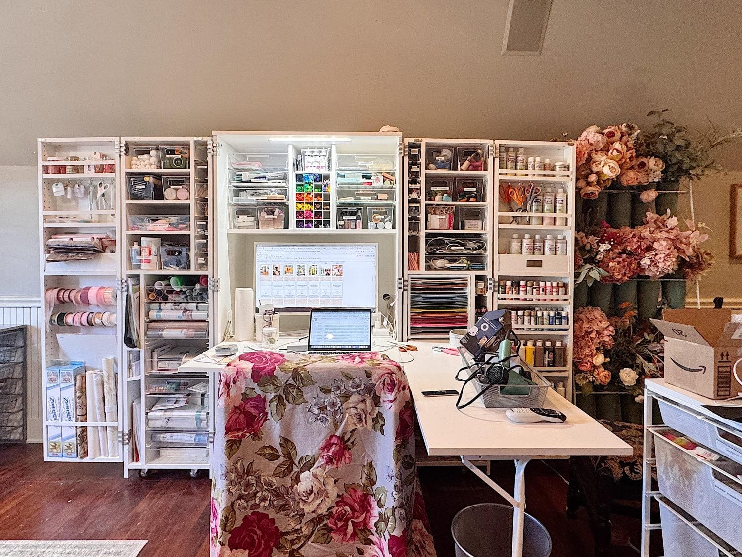 A craft workspace with open shelves filled with art supplies, a laptop on the desk, and artificial flower arrangements on the right.