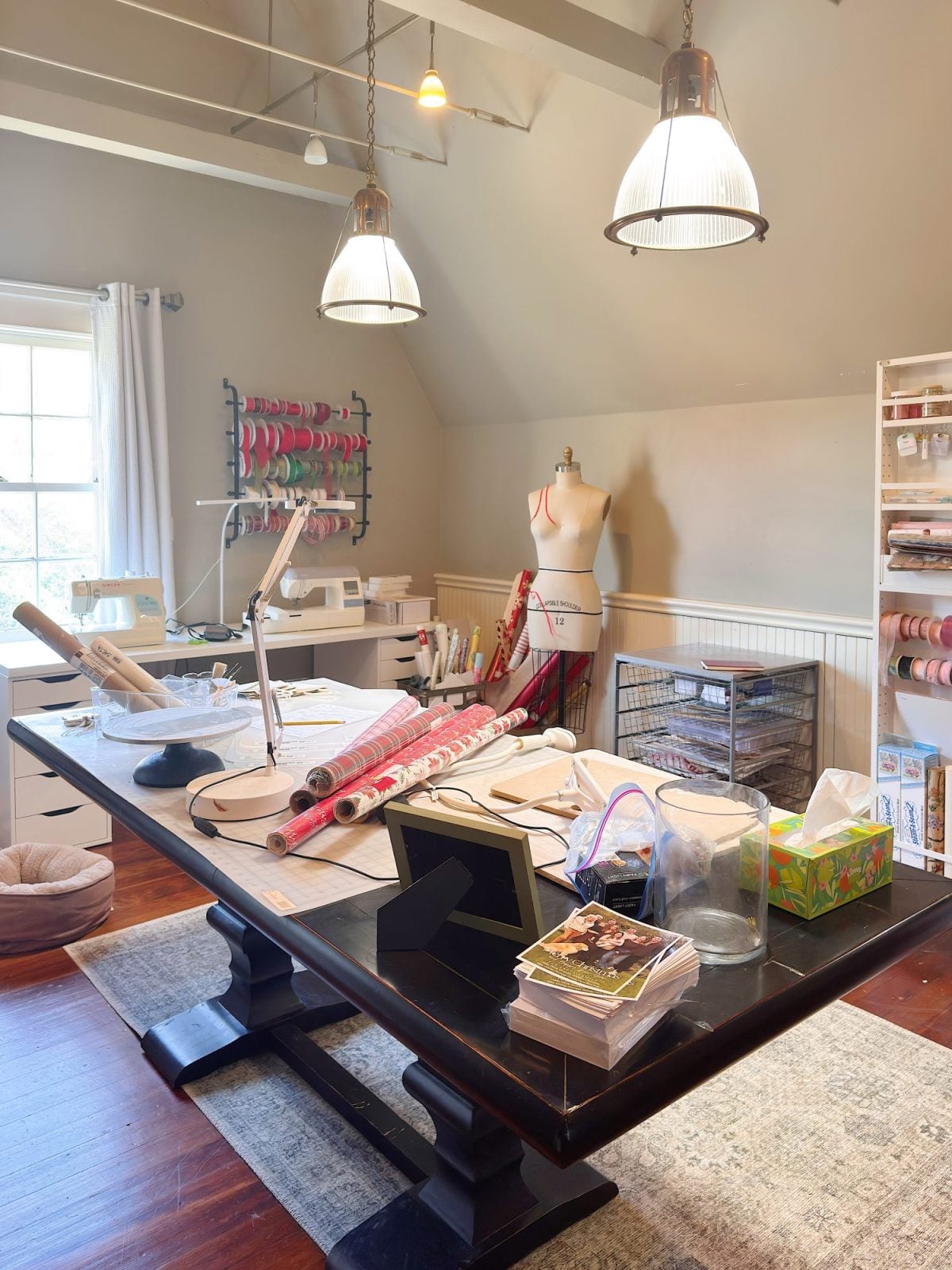 A craft room with a large table covered in wrapping paper rolls, tools, fabric, and supplies, with shelves, a dress form, and organized ribbons in the background.